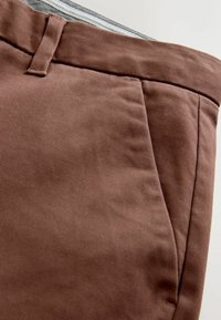 Braune Baumwollhosen mit glatter Textur, ausgestattet mit Seitentaschen und einem ordentlichen Bund. Eine Nahaufnahme hebt die Webart des Stoffes hervor.