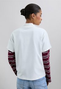 Top corto blanco con cuello, superpuesto a una camiseta de manga larga color marrón con rayas horizontales blancas, combinado con jeans de mezclilla azul claro.