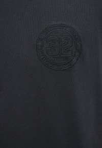 Schwarzes, texturiertes Stoff-T-Shirt mit einem kreisförmigen, geprägten Logo in der Mitte, auf dem die Zahl "32" zu sehen ist, umgeben von dem Markennamen und dem Gründungsdatum.