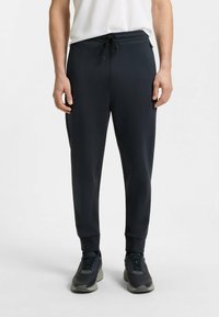 Navy jogger-housut, jotka on valmistettu sileästä kankaasta, sisältävät joustovyötärön ja nauhan, sivutaskut ja kapenevat hihnat.