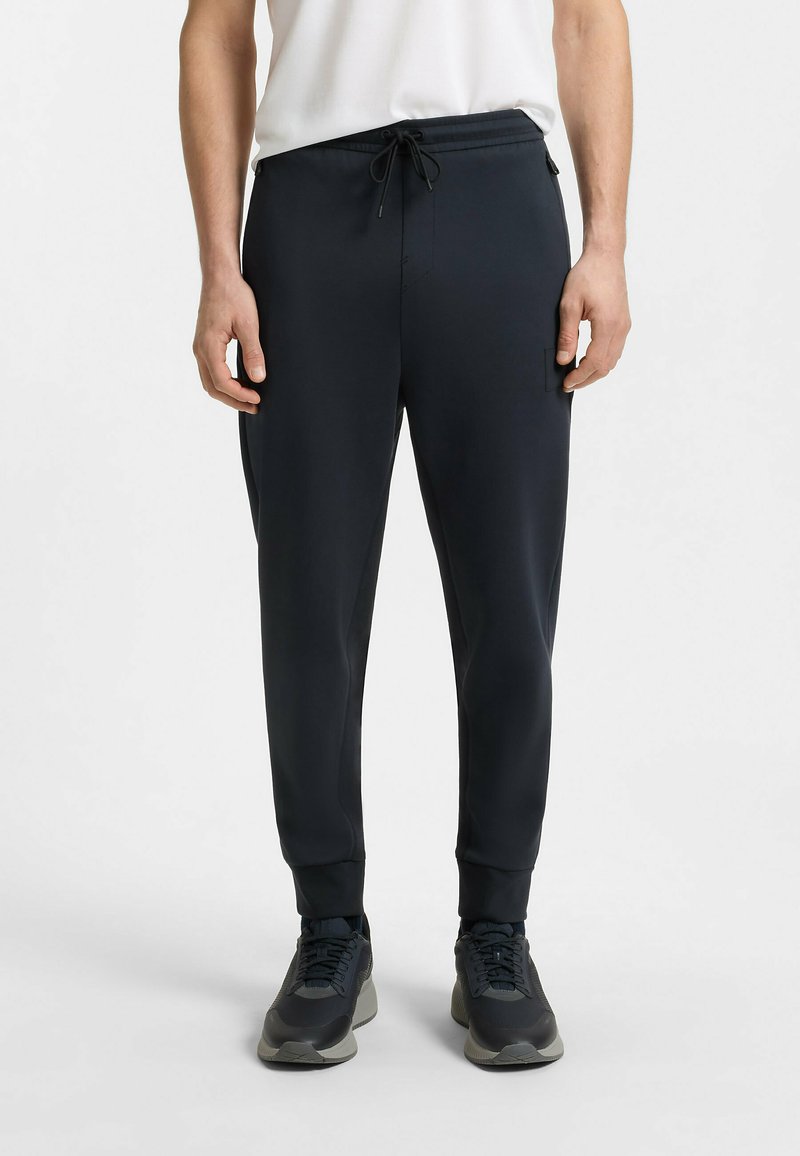 Navy jogger-housut, jotka on valmistettu sileästä kankaasta, sisältävät joustovyötärön ja nauhan, sivutaskut ja kapenevat hihnat.