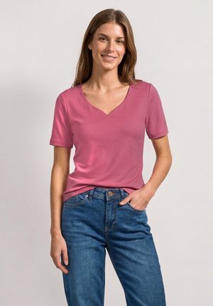 Młoda kobieta o długich brązowych włosach, ubraną w różowy t-shirt z krótkim rękawem i dekoltem w serek oraz niebieskie dżinsy, uśmiechającą się z jedną ręką w kieszeni.