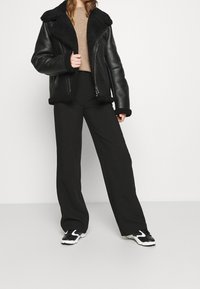 Czarna skórzana kurtka z kołnierzem z owczej wełny, w zestawieniu z beżowym swetrem i szerokimi czarnymi spodniami. Model nosi czarno-białe sneakersy.