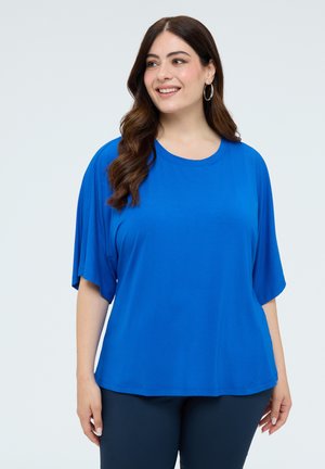 Lachende vrouw met lang bruin haar met hoepeloorbellen, een felblauwe losse korte mouw top en donkere broek tegen een effen achtergrond.