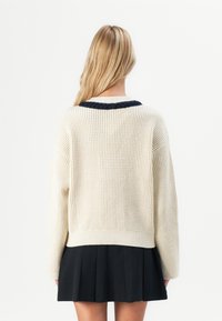 Femeie cu păr lung, blond, îmbrăcată cu un pulover tricotat crem cu margine bleu închis și o fustă plisată neagră, stând pe un fundal alb.