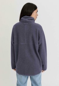 Pullover in pile viola con collo alto, spalle scese, una cucitura sulla schiena e una vestibilità rilassata, abbinato a jeans azzurri chiari.
