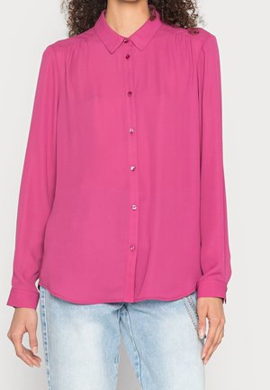 Kvinde iført en langærmet, knap-op lyserød skjorte med krave, kombineret med lyseblå jeans.