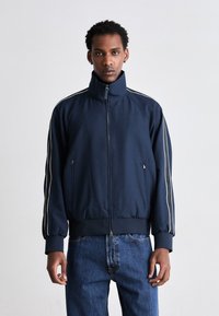 Jachetă navy cu fermoar, cu guler înalt, dungi laterale albe și mansete ribbate. Țesătură netedă și design ajustat, asortată cu blugi din denim albastru.