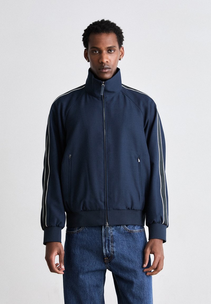 Jachetă navy cu fermoar, cu guler înalt, dungi laterale albe și mansete ribbate. Țesătură netedă și design ajustat, asortată cu blugi din denim albastru.