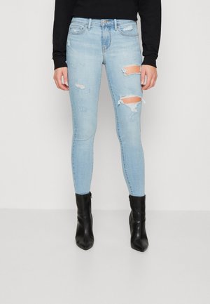 Femme portant un jean skinny bleu clair déchiré, des bottines noires à talons, et un haut noir à manches longues, debout sur un sol gris contre un mur blanc.
