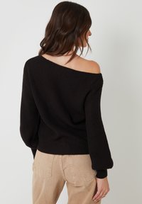 Zwarte geribbelde gebreide sweater met een off-shoulder ontwerp en ballonmouwen, gecombineerd met lichtbeige corduroybroek.