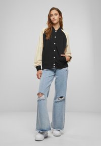 Svart og krem oversized bomberjakke med ribbestrikkede mansjetter og seks hvite knapper, kombinert med lyseblå ripped flare-jeans og hvite joggesko.