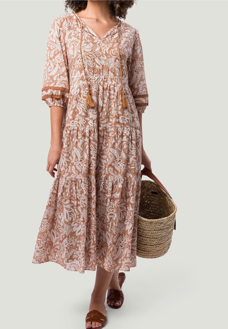 Femme dans une robe cachemire marron et blanche, tenant un sac panier tissé, portant des sandales marron, debout devant un fond uni.