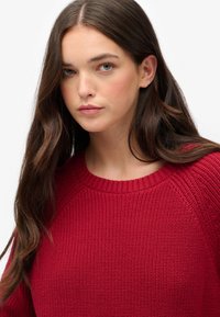 Maglione rosso lavorato a maglia con scollatura rotonda a coste e maniche raglan. Presenta un tessuto testurizzato con uno stile aderente.