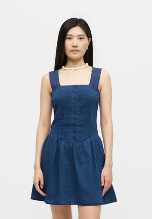 Γυναίκα με ίσια μαύρα μαλλιά που φορά αμάνικο μπλε φόρεμα με κουμπιά και δαντελωτούς ιμάντες και λευκό κολιέ με χάντρες σε λευκό φόντο.