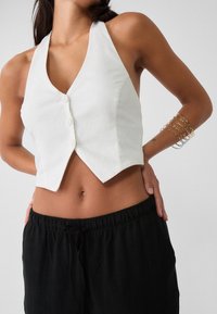 Weißes, kurz geschnittenes Halter-Top aus Leinen mit tiefem V-Ausschnitt, fünf Knöpfen vorne und einem strukturierten Design. Kombiniert mit schwarzen Hosen mit Kordelzug.