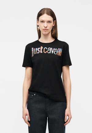 Nő egyenes barna hajjal, nagy arany cseppalakú fülbevalókat visel, valamint fekete pólót színes "Just Cavalli" felirattal, sötét farmerrel párosítva.