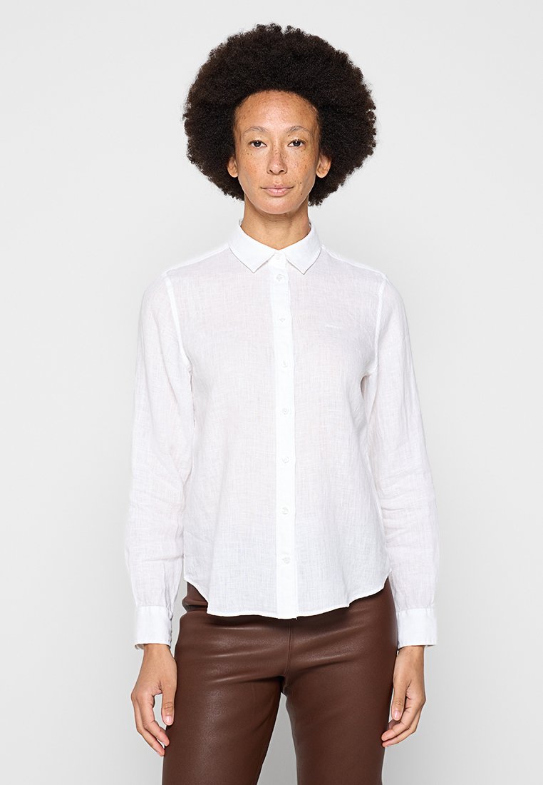 Biała lniana koszula z długim rękawem, z guzikami z przodu, kołnierzykiem i luźnym krojem. Połączona z brązowymi dopasowanymi spodniami, widoczna gładka tekstura.