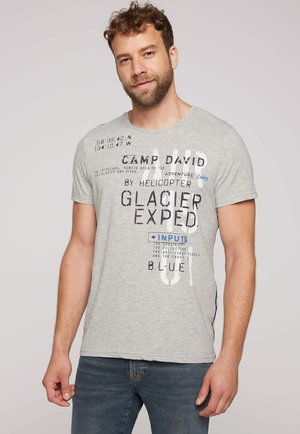 Uomo con capelli ricci e barba che indossa una t-shirt grigio chiaro con scritte nere e blu e jeans blu scuro, in piedi davanti a uno sfondo semplice.