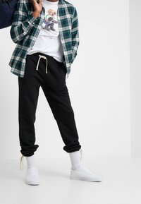 Czarne dresy z troczkami, białe sneakersy, zielona i granatowa koszula w kratę oraz biała koszulka z grafiką przedstawiającą niedźwiedzia.