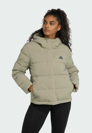 Žena s kudrnatými vlasy, která nosí světle šedou prošívanou bundu s kapucí, ruce v kapsách, dívá se stranou, stojí před jednobarevným pozadím.