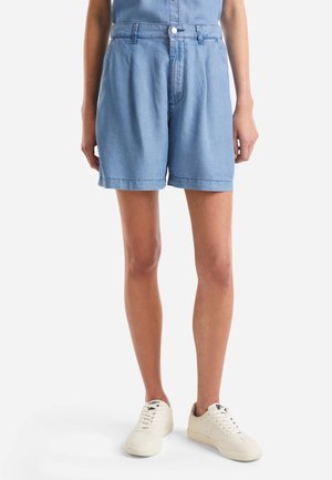 Person iført lyseblå denimshorts og hvide sneakers står mod en ensfarvet hvid baggrund.