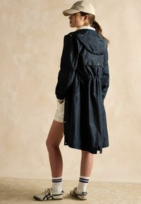 Tumši zils vēja jaka ar sašaurinātu vidukli, kapuci un ar rāvējslēdzējiem aizveramām kabatām, apvienota ar bežām šortiem un sporta apaviem. Cepure ar vienkāršu dizainu.