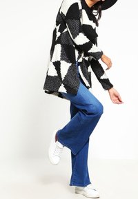 Maglione lavorato a maglia oversize bianco e nero con motivo geometrico, abbinato a jeans a zampa d'elefante blu e sneakers bianche. Modella in posa di profilo.