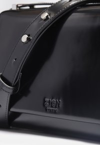 Black patent leather bag with a smooth texture, silver hardware accents, and a logo embossed on the side. Strap shown partially.