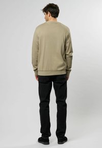 Camicia beige a maniche lunghe con scollo rotondo, abbinata a pantaloni e scarpe neri. Il tessuto appare liscio con dettagli a coste sui polsini e sull'orlo.
