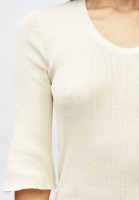 Haut en tricot côtelé beige clair avec un décolleté arrondi et des manches bouffantes trois-quarts. Présente une texture douce et un léger extensibilité.