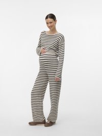 Έγκυος γυναίκα με μπεζ και μαύρη ριγέ άνετη ενδυμασία, όρθια, χαμογελά και κρατά την κοιλιά της με το ένα χέρι.