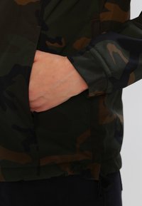 Camouflagejakke med en blanding af mørkegrønne og brune farver, med en lynlåslomme og et blødt, tekstureret stof.