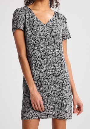 Frau trägt ein kurzärmliges schwarz-weißes Kleid mit Blattmuster und V-Ausschnitt, steht vor einem einfarbigen Hintergrund.