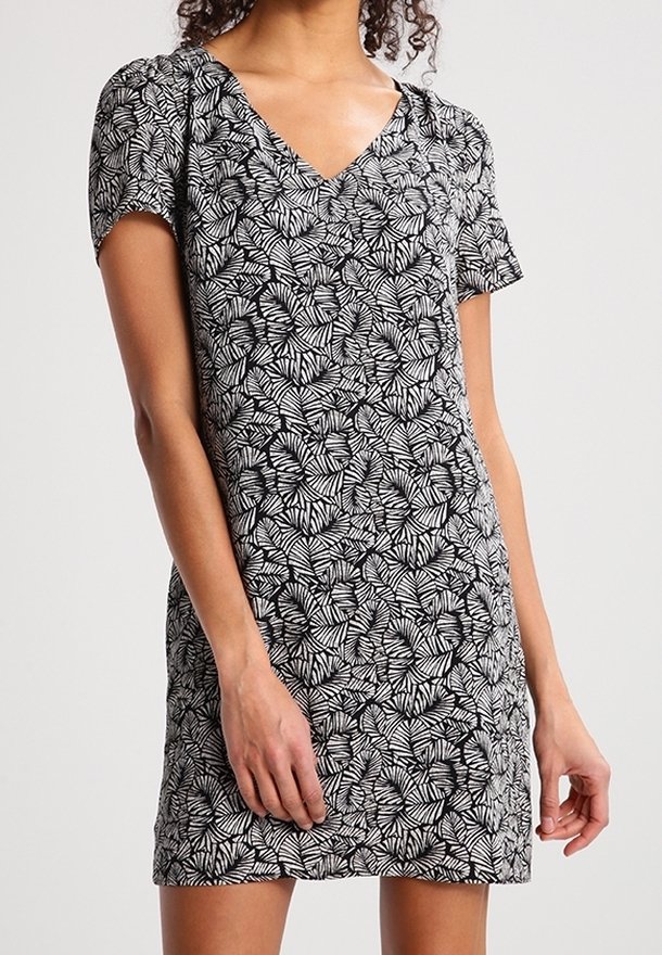 Femme portant une robe noire et blanche à manches courtes avec un motif de feuilles et un décolleté en V, debout devant un fond uni.