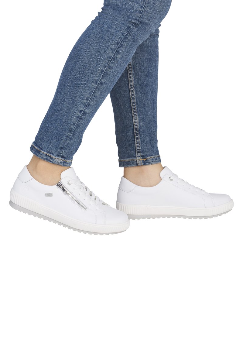 Sneakers bianche con lacci e cerniera laterale indossate con jeans skinny blu, mostrate a metà passo su sfondo bianco.