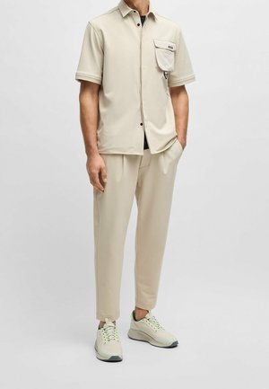 Camisa de manga corta beige con un bolsillo, combinada con pantalones beige holgados y zapatillas de color claro con acentos verdes. Textura suave.