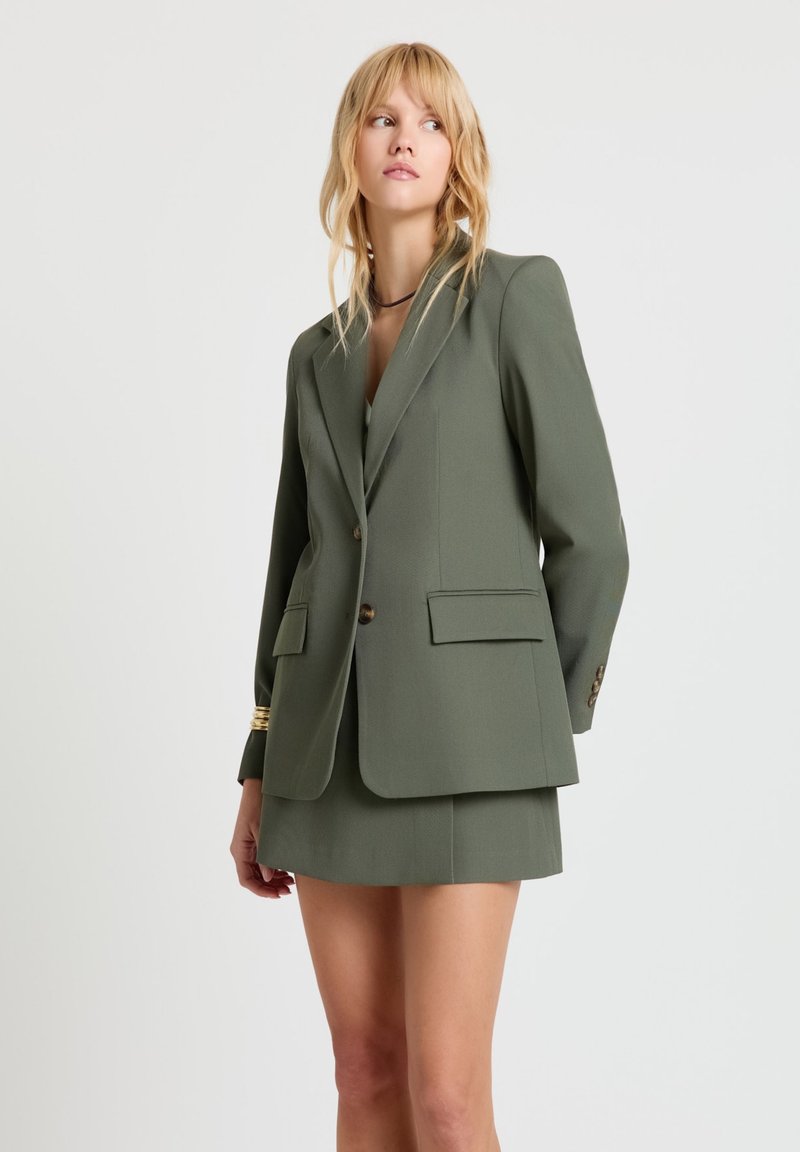 Blonde Frau, die einen olivegrünen Blazer und einen passenden Minirock trägt, steht mit den Händen hinter dem Rücken und blickt zur Seite vor weißem Hintergrund.