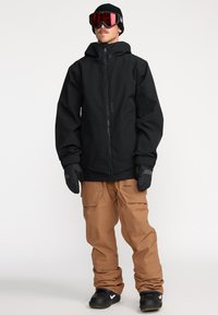 Veste imperméable noire avec capuche, associée à un pantalon de neige ample marron. Comprend des gants et des lunettes de ski avec un verre rouge.