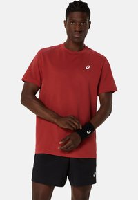 Červené sportovní tričko s krátkým rukávem a kulatým výstřihem, s detaily loga, spárované s černými šortkami a černým zápěstním páskem. Hladká textura.