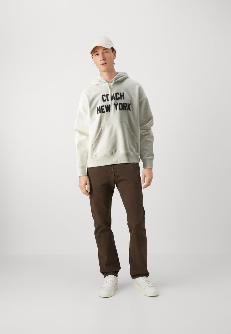 Lichtgrijze hoodie met "COACH NEW YORK" in vet zwart, bruine broek en witte sneakers. De hoodie heeft een voorzak en een capuchon met trekkoord.