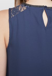 Top senza maniche blu navy con dettagli in pizzo sulle spalle, apertura a chiave sul retro e tessuto testurizzato. Presenta un bottone decorativo al collo.
