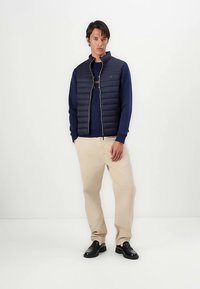 Colete acolchoado azul-marinho com fecho de correr, usado sobre um suéter azul-marinho, combinado com calças creme e sapatos pretos, contra um fundo branco liso.