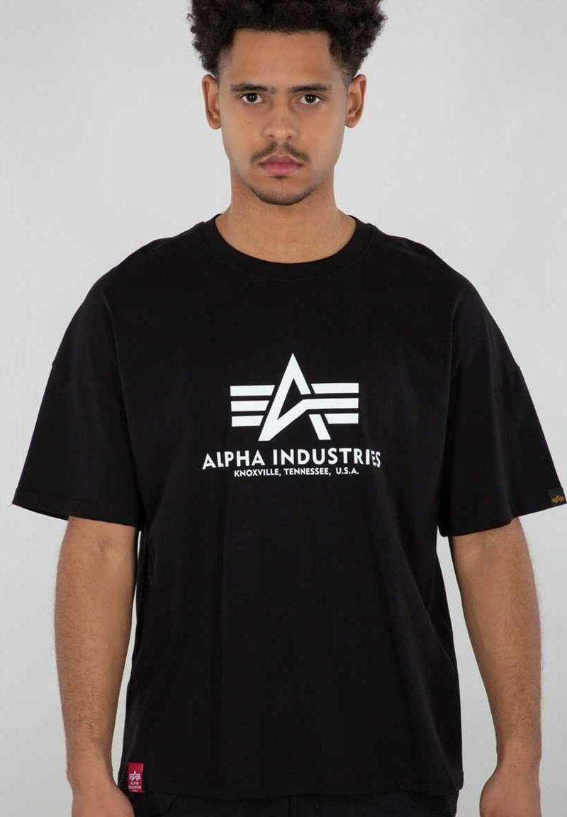 Ung mand med kort krøllet hår iført en sort Alpha Industries T-shirt med hvidt logo og tekst, stående mod en ensartet lys baggrund.