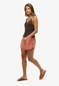 Top sin mangas de punto marrón con tirantes finos, combinado con pantalones cortos rosas claros. La modelo lleva sandalias marrones de tipo slide. Un atuendo simple y casual.