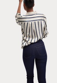 Blouse à manches longues rayée en crème et bleu marine, associée à un jean bleu foncé. La blouse possède un col et une coupe décontractée, avec des plis visibles dans le dos.