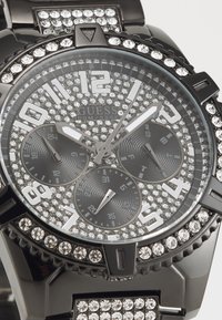 Svart klocka med en juvelbelagd urtavla, silverfärgade timmarkörer och tre sub-dialer. Glansig rem prydd med rhinestones.