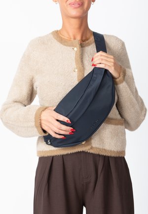 Bolso bandolera azul marino hecho de tela suave, con cierre de cremallera y marca discreta. Modelo sostiene el bolso mientras lleva un cárdigan beige.