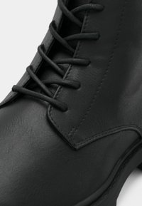 Bottine en cuir noir à texture lisse, avec un laçage complexe et un design structuré. Détails cousus le long des bords.