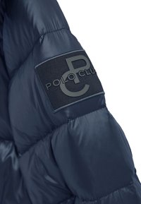 Marinblå vadderad jacka med quiltad textur och en stor svart patch på ärmen med grå texten "POLO CLUB" och logotyp.